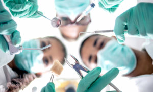 Looking up into 3 oral surgeons with turquoise mouth masks holding dental surgery tools