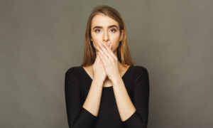 Blonde woman wearing a black shirt covers her mouth with her hands because she is embarrassed about her halitosis
