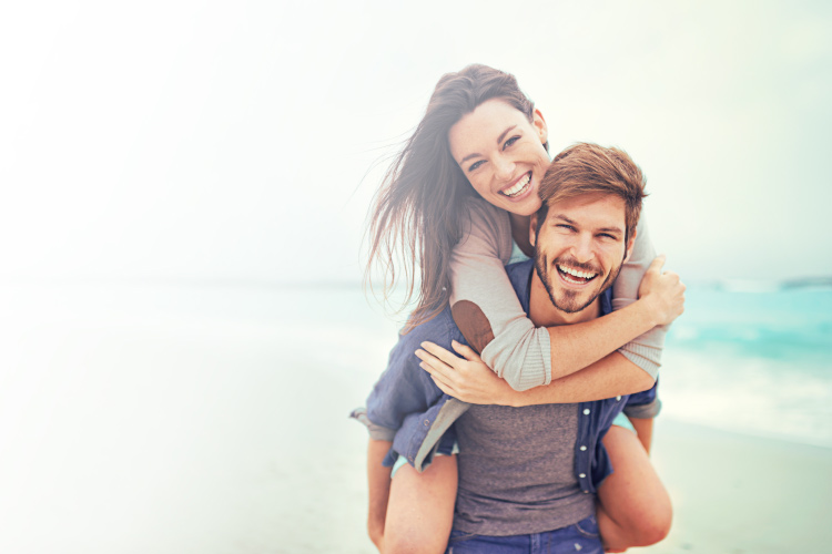 Brunette man gives a piggy back ride to a brunette woman while smiling on a beach