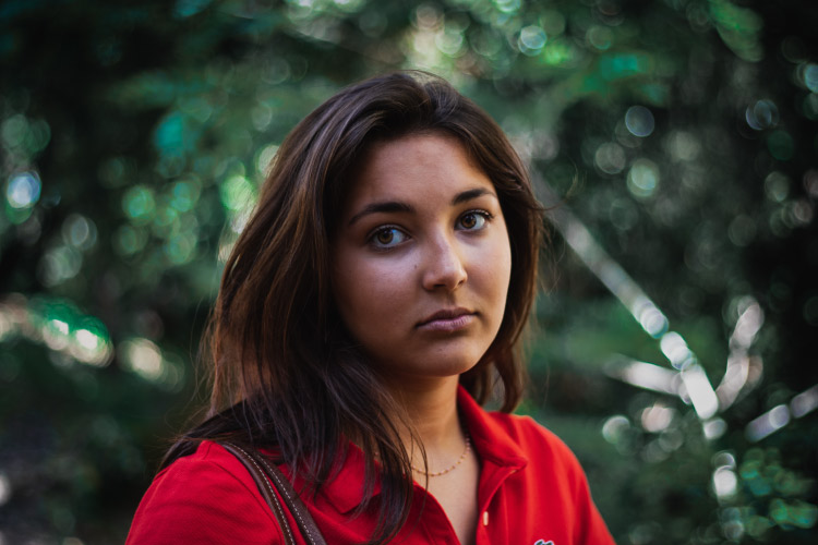 Brunette woman with dental anxiety wears a red blouse outside near green foliage