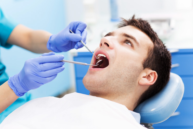 Brunette man sits in a dental chair with a white bib as a hygienist examines his mouth with dental tools