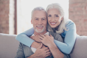 A white-haired husband and wife smile with dentures as they embrace on the couch