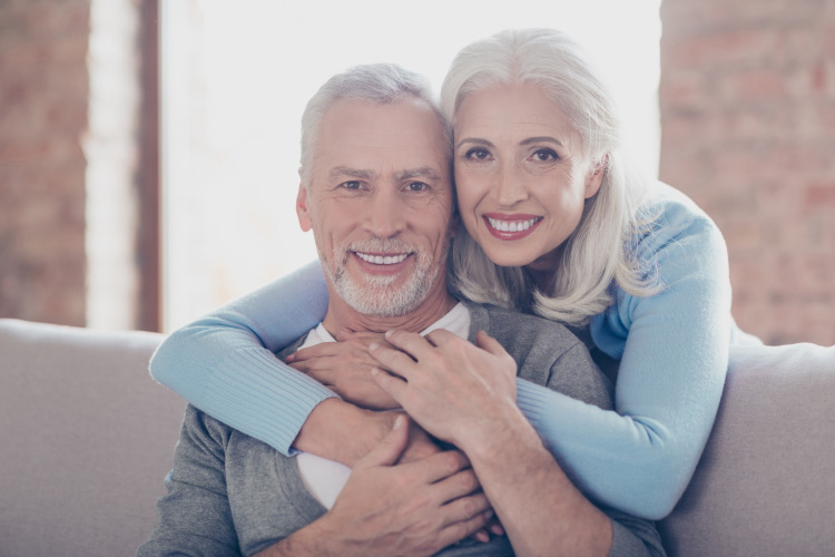 A white-haired husband and wife smile with dentures as they embrace on the couch