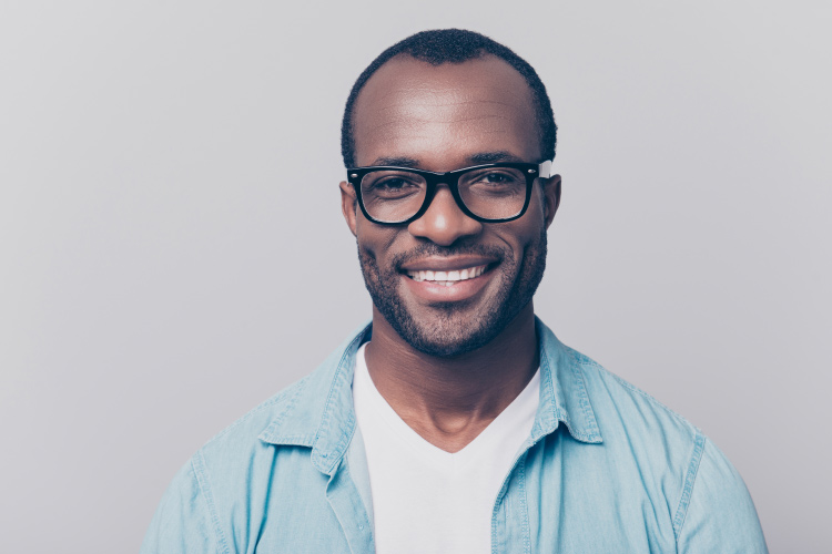 Dark-haired man with black glasses and a sky blue shirt smiles with his dental crowns