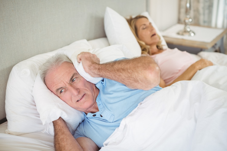 An older man in bed holds a pillow over his ear while his wife with sleep apnea snores loudly next to him