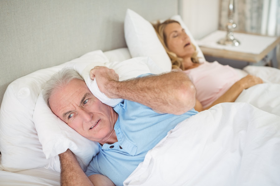 Snoring woman with sleep apnea next to her husband who has a pillow over his ears.